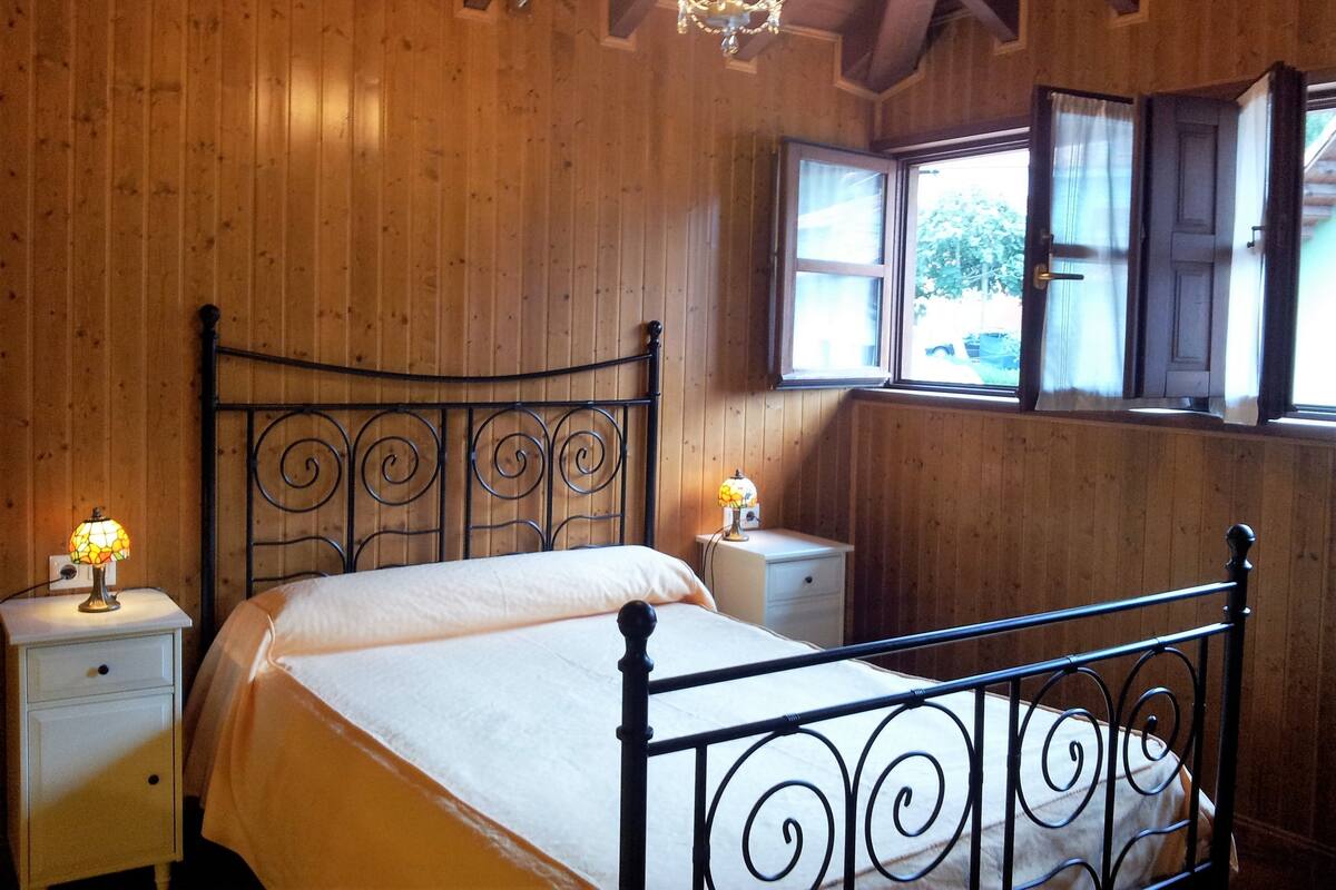 Un dormitorio con paneles de madera, una estructura de cama metálica, dos mesitas de noche con lámparas y una ventana con cortinas.