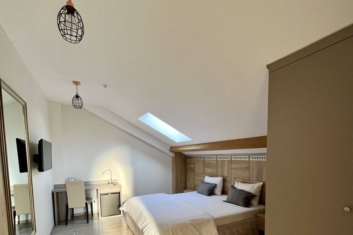 A modern bedroom with a large bed, a desk, and a chair. There is a skylight above the bed and a wardrobe on the right.