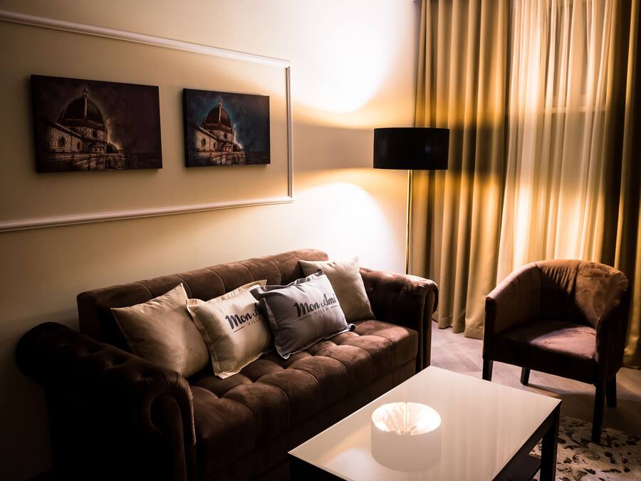 A living room with a brown sofa, two pillows, a white coffee table, a lamp, and a chair. There are two framed artworks on the wall.