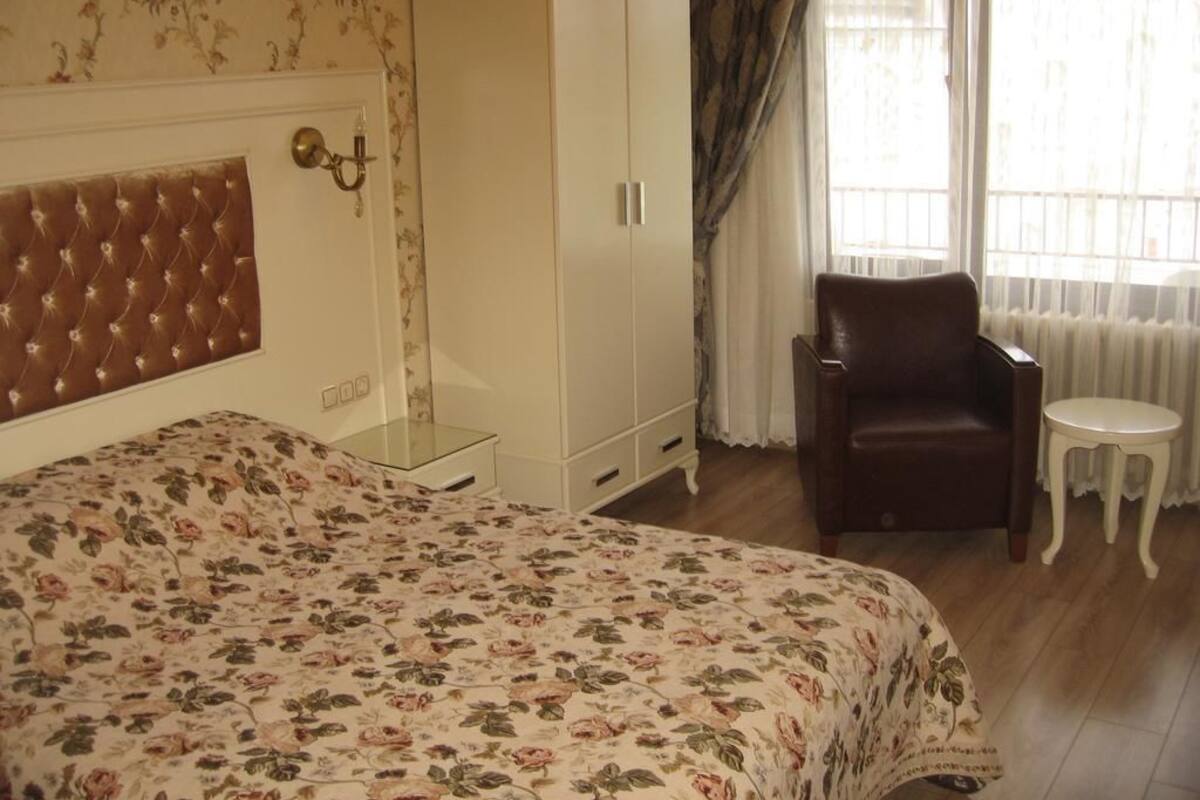 Un dormitorio con colcha estampada, cabecera capitonada, armario blanco, sillón de cuero y una mesita redonda.