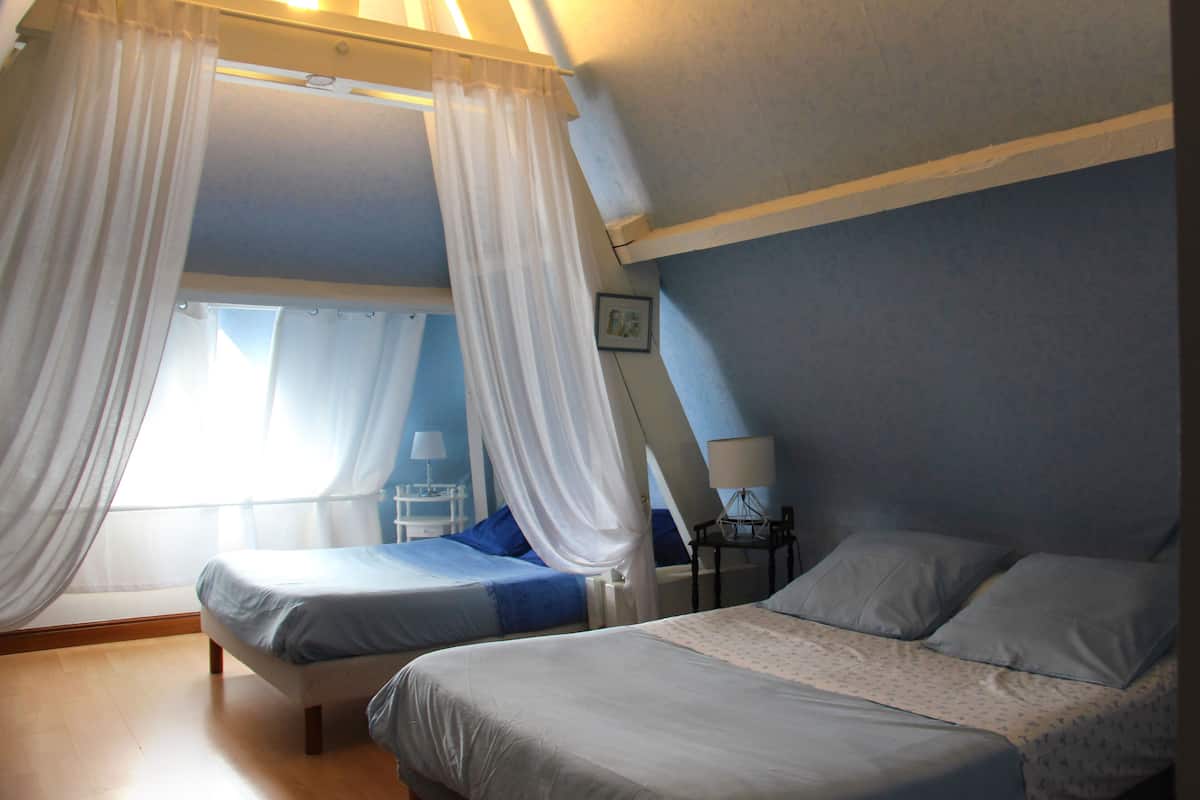 Une chambre avec deux lits, une fenêtre avec des rideaux légers, une table de chevet avec une lampe et un tableau accroché au mur.