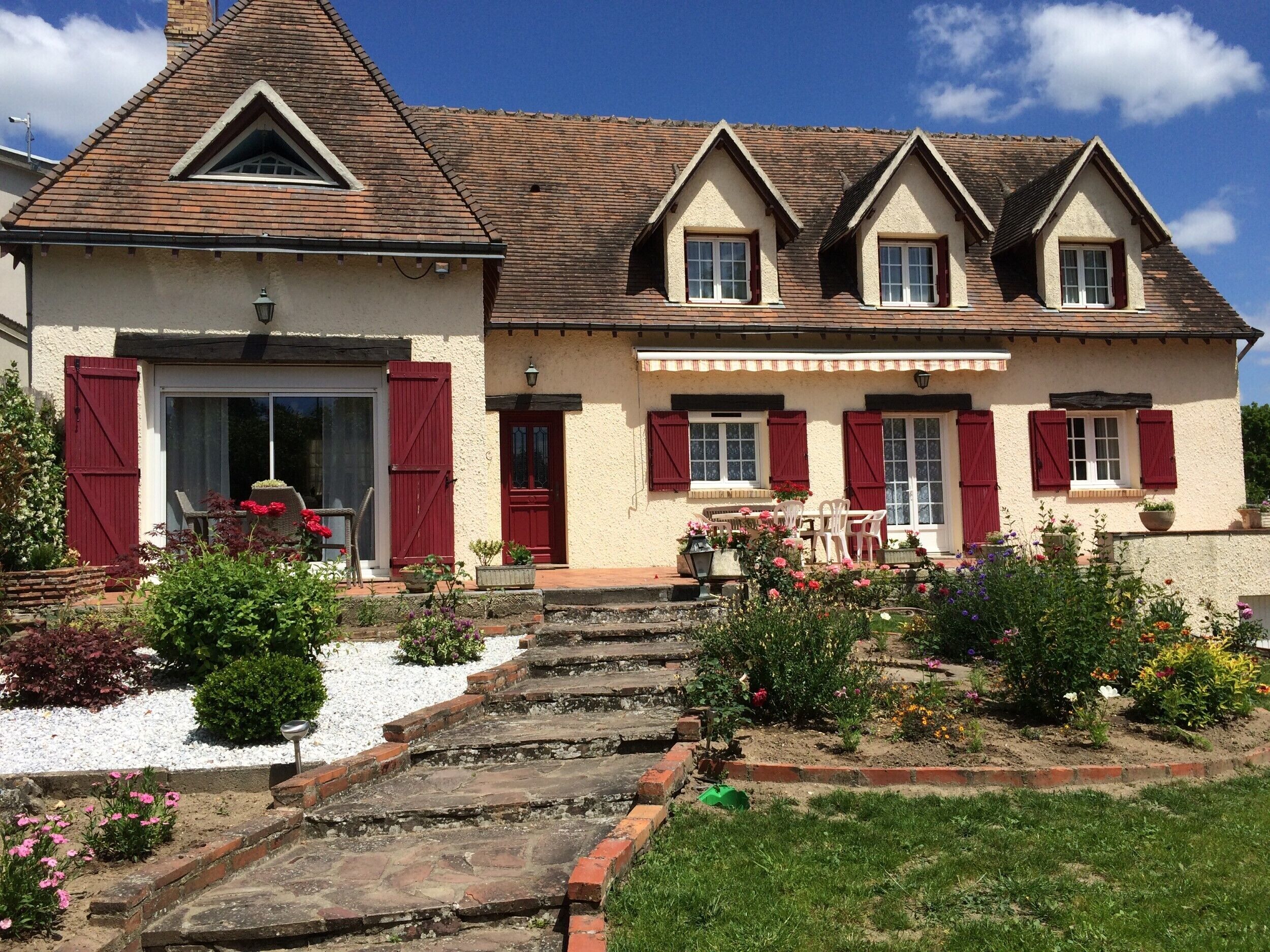 Uma casa de dois andares com telhado inclinado, entrada frontal com porta de vidro e um jardim com diversas plantas e flores.