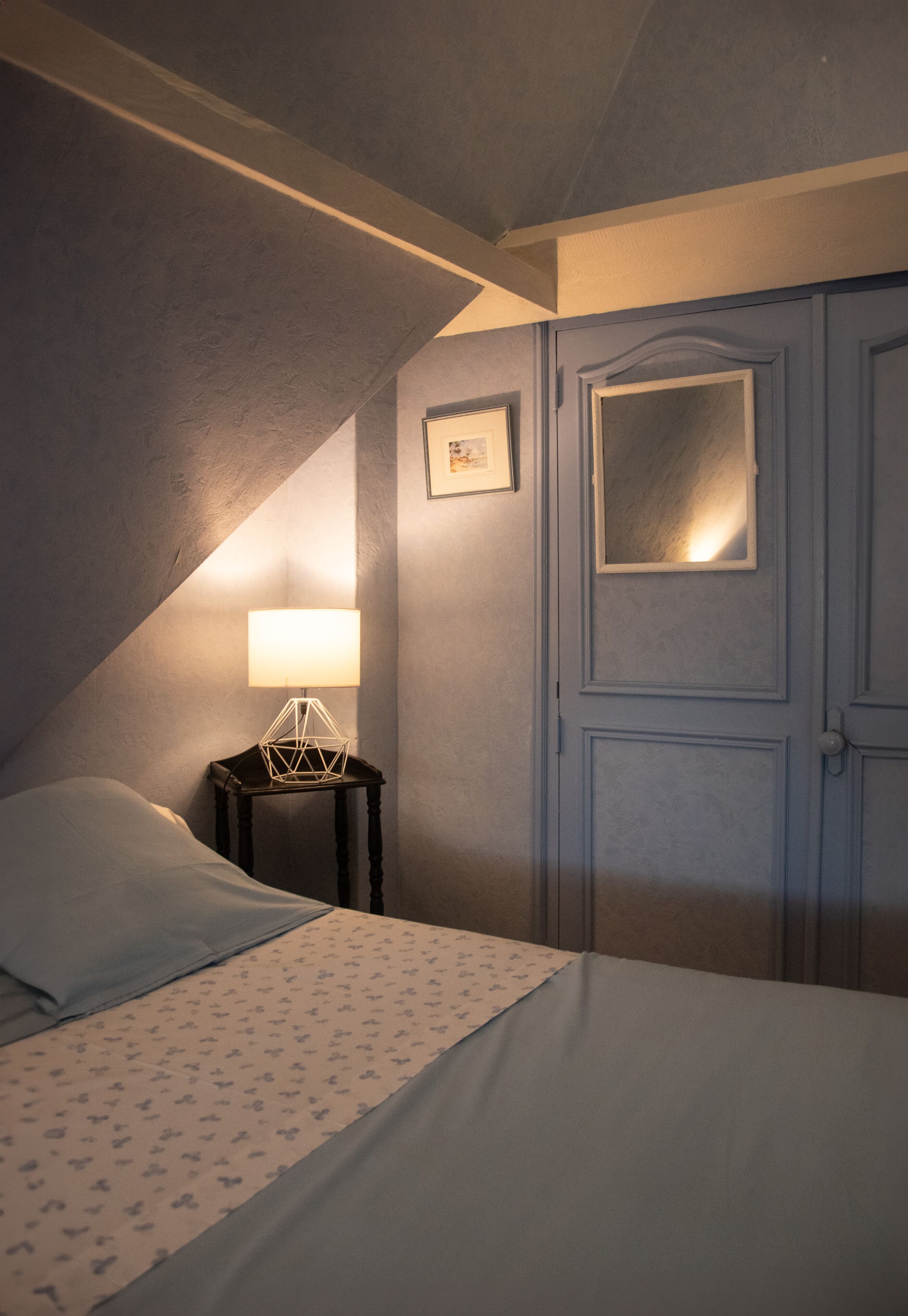 A bedroom with a bed, a nightstand with a lamp, a framed picture on the wall, and a door.