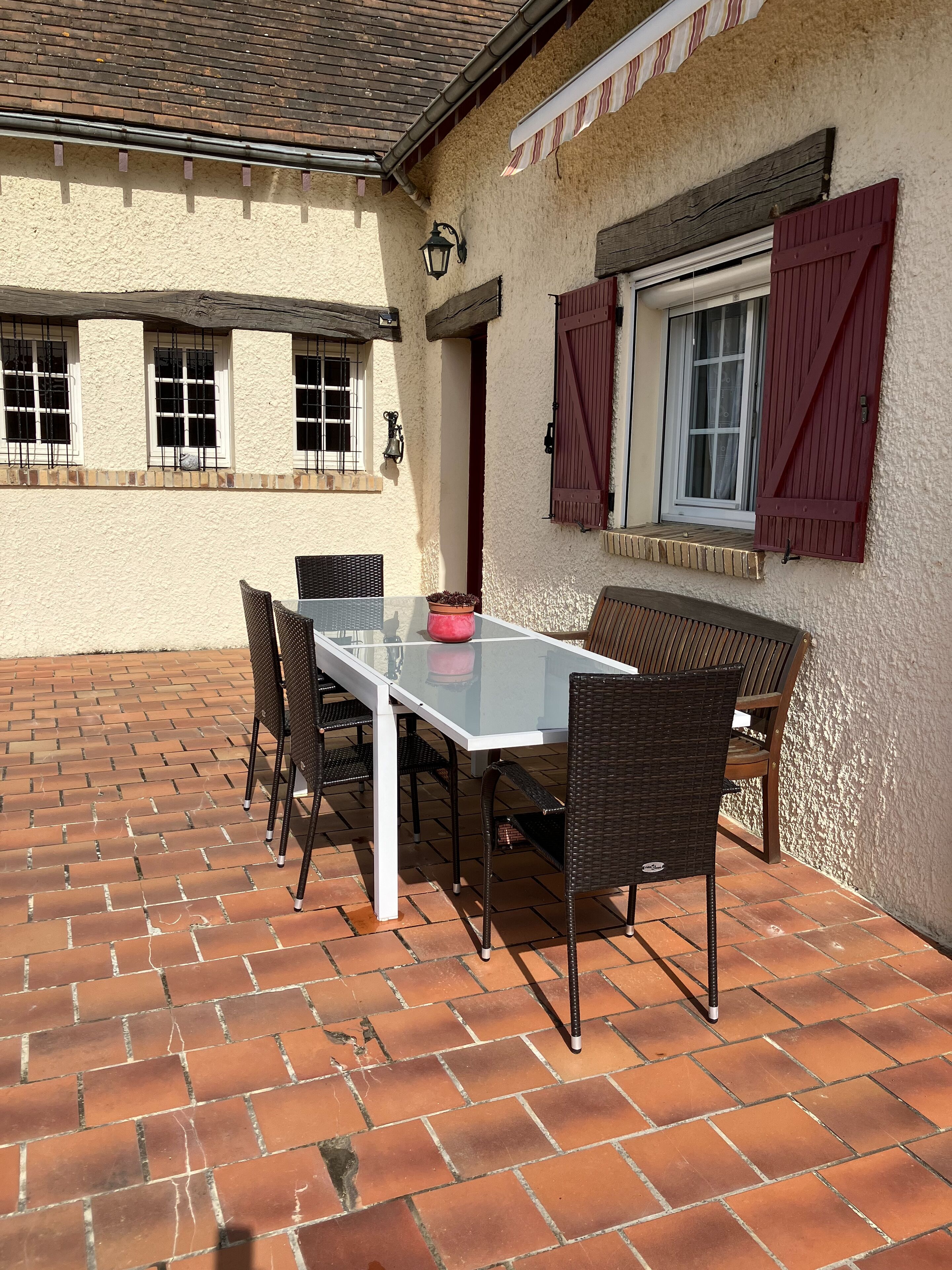 Une terrasse extérieure avec une table et des chaises, un sol en briques et un bâtiment doté de fenêtres et de volets.