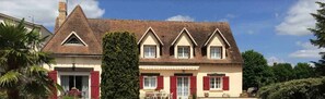 Une maison à deux étages, dotée d’un toit à forte pente, de lucarnes et d’un porche à l’avant, avec une table et des chaises.