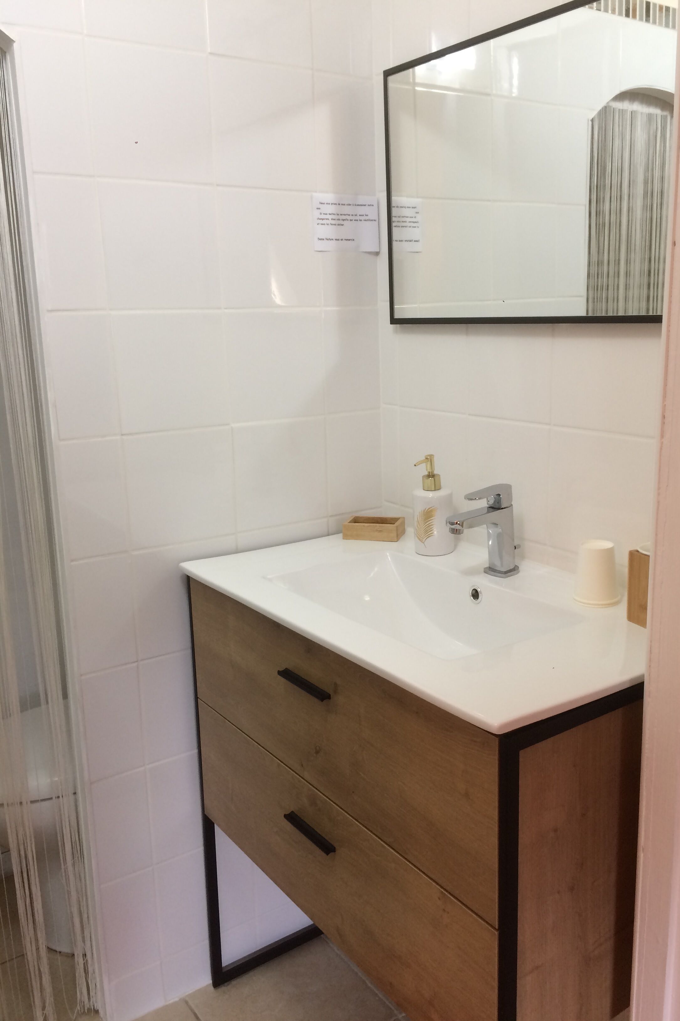 Une salle de bain carrelée de carreaux blancs, avec un meuble-lavabo en bois doté d’un miroir, un évier et des toilettes.