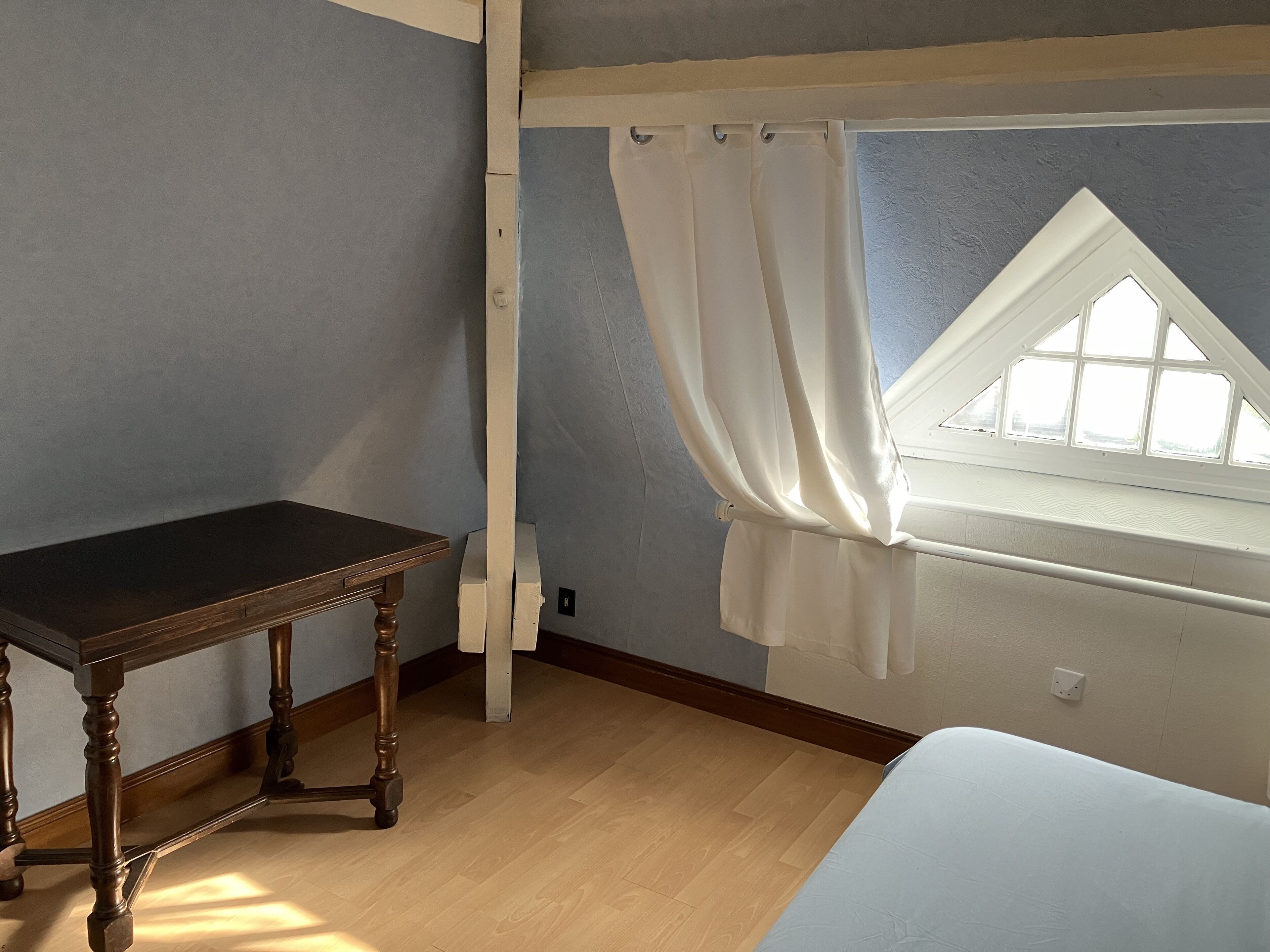 A room with a wooden table, a window with a white curtain, and a bed.