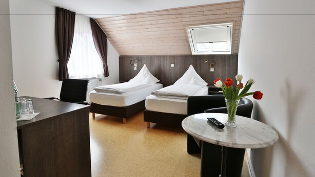 A hotel room with two beds, a desk, a chair, a round table with a vase of flowers, and a TV mounted on the wall. - Hotel Gasthof Roessle (Rottenburg)