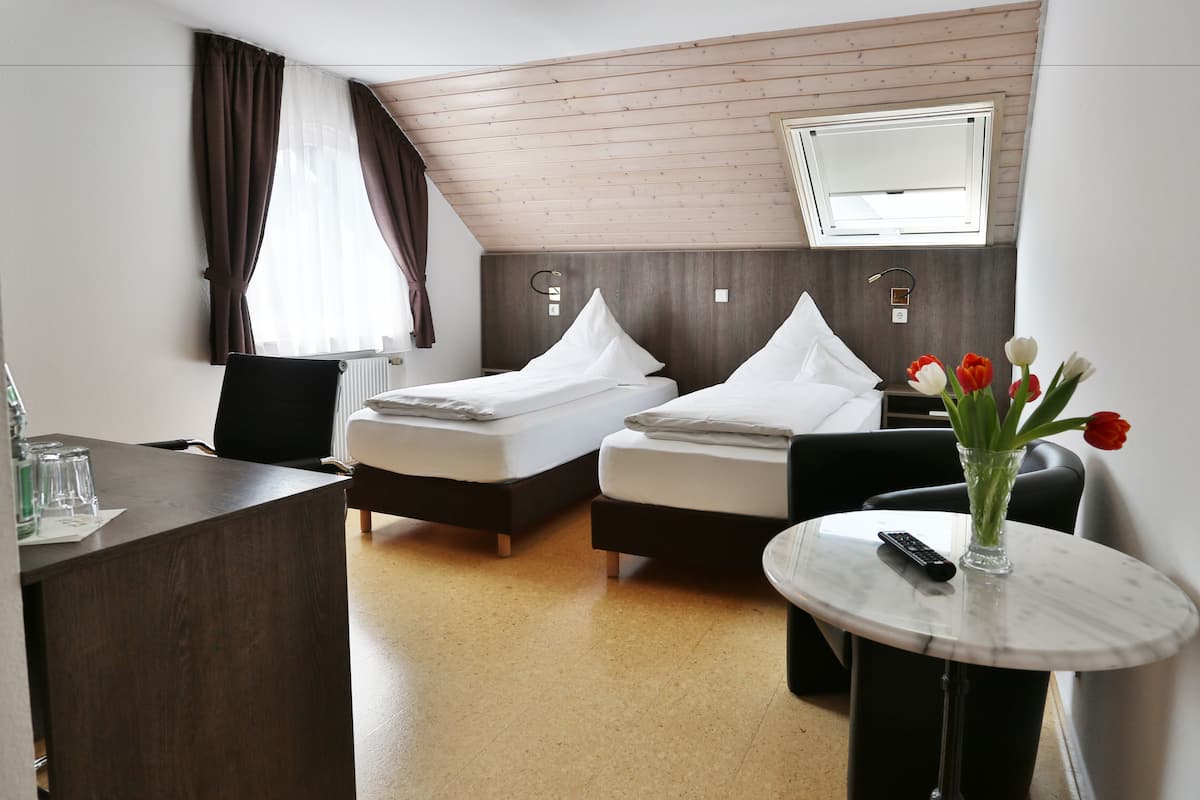Habitación de hotel con dos camas, un escritorio, una silla, una mesa redonda con un jarrón de flores y un televisor montado en la pared.
