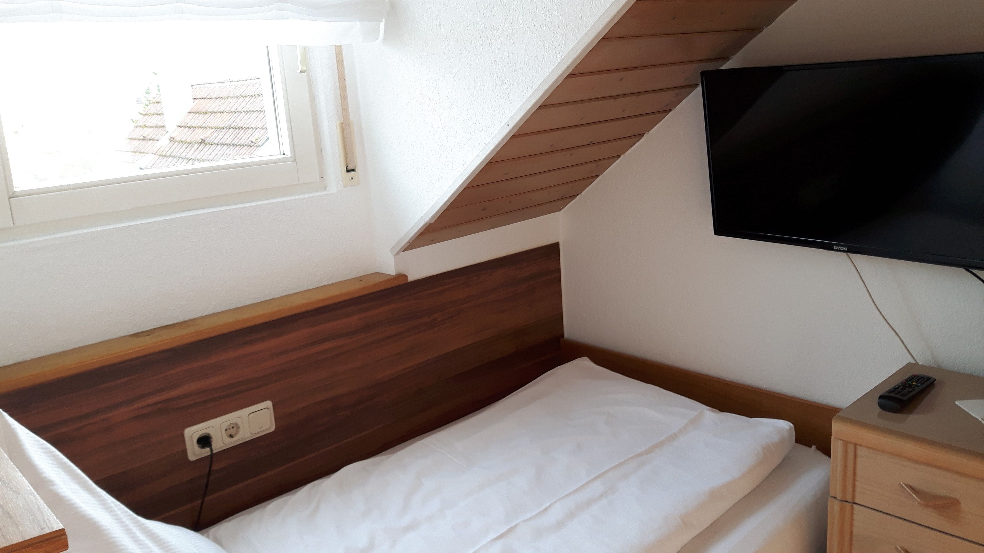A bedroom with a wooden headboard, a bed with white bedding, a television mounted on the wall, and a window with a view of the outside.