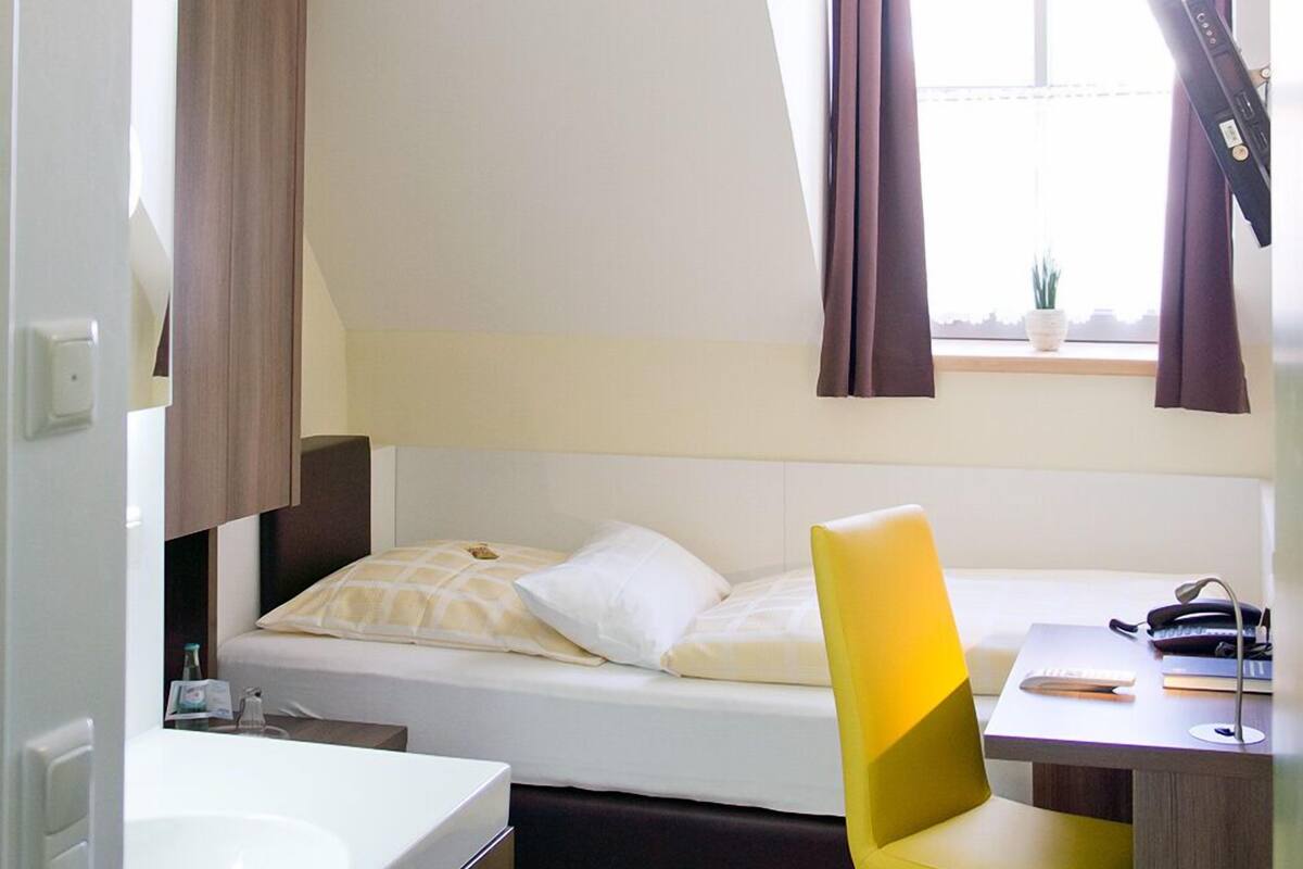 Een slaapkamer met een bed, bureau, stoel, raam met gordijnen en een badkamer met wastafel.