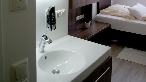 Een moderne hotelkamer met een wit wastafel, een spiegel, een klein rond spiegeltje, een bed met witte lakens en een raam met gordijnen.