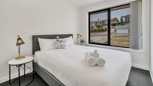 A hotel room with a large bed, a small bedside table with a lamp, a window with a view of the outside, and a neatly made bed with towels. - Youngtown Executive Apartments (Youngtown)