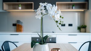 A modern kitchen with white cabinets, a wooden table, a white vase with white flowers, and a copper mug. - Youngtown Executive Apartments (Youngtown)