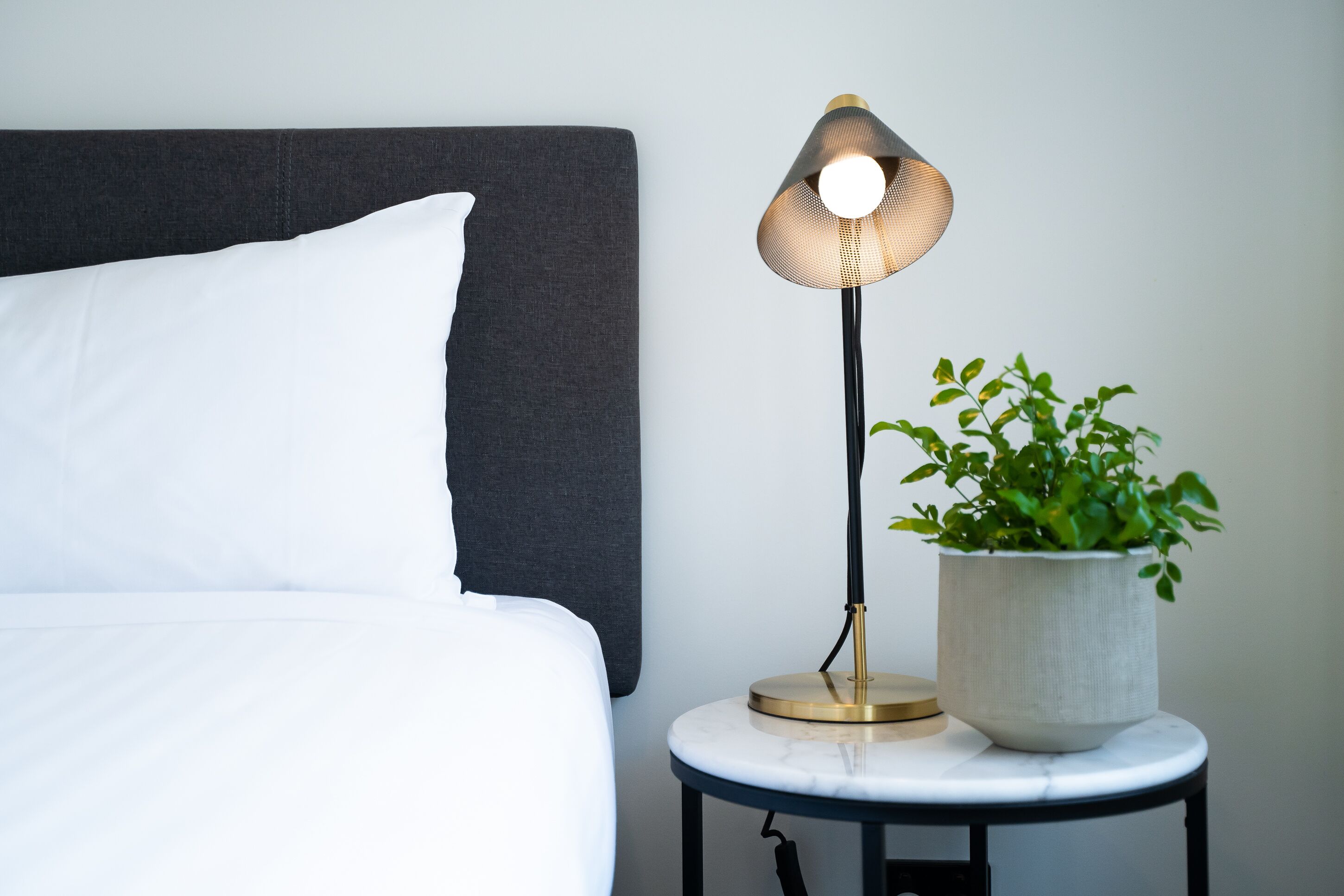 A neatly made bed with white bedding, a dark headboard, a modern table lamp with a gold base, and a potted plant on the side table.