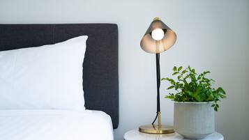A neatly made bed with white bedding, a dark headboard, a modern table lamp with a gold base, and a potted plant on the side table.