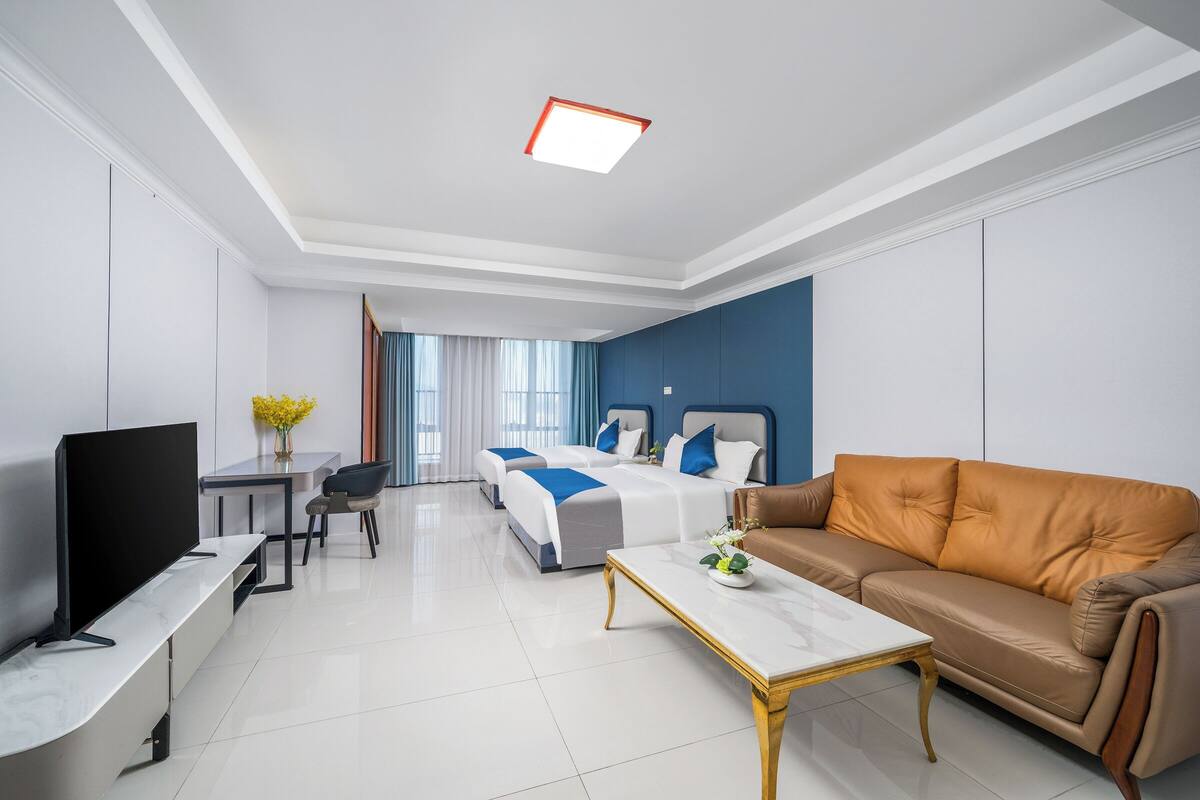 A modern hotel room with a flat-screen TV, a sofa, a bed with blue bedding, a bedside table, and a window with curtains.