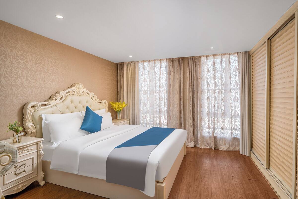 A hotel room with a large bed, a nightstand, a vase with flowers, and a window with curtains.