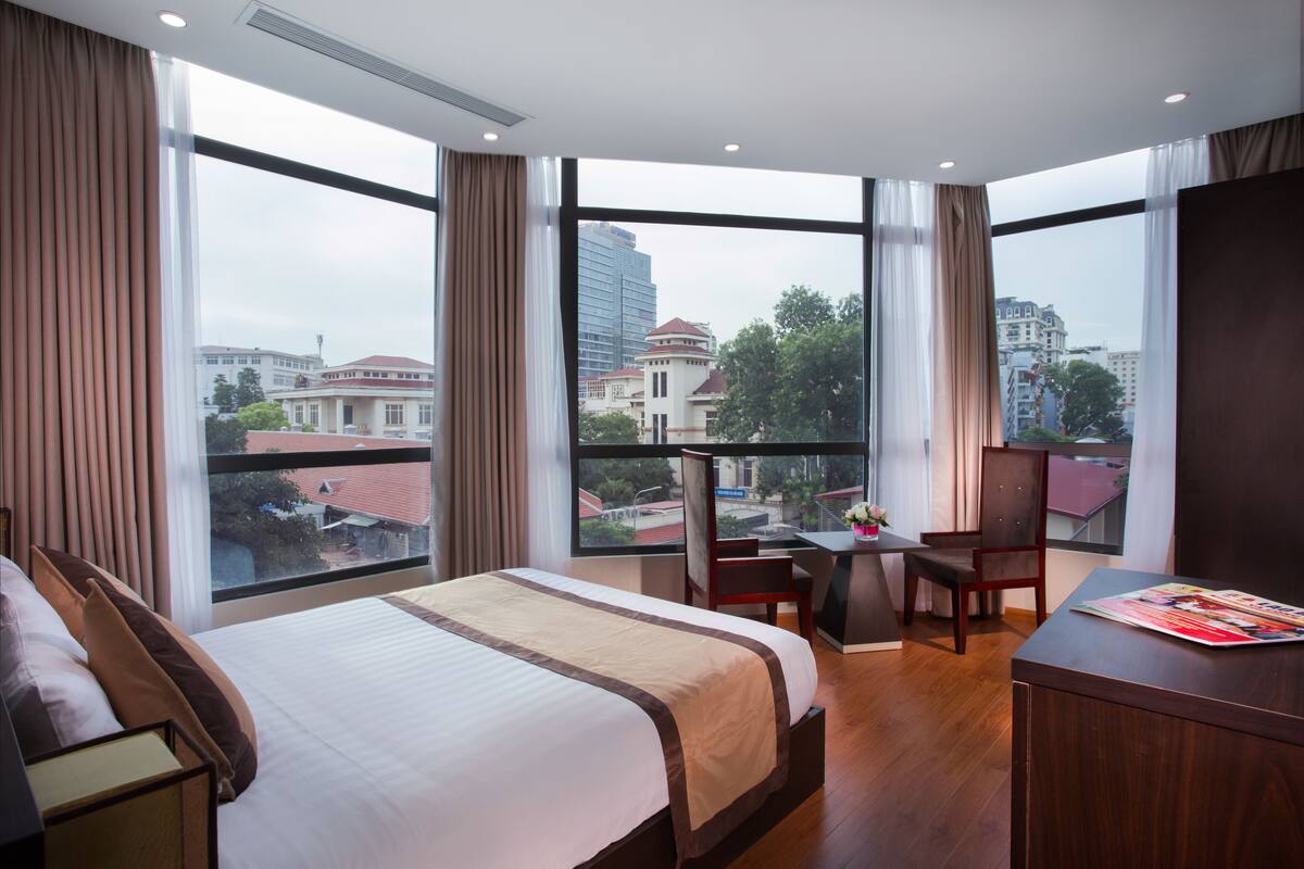 A hotel room with a large bed, a desk, and a chair, a view of the city through large windows, and a balcony.