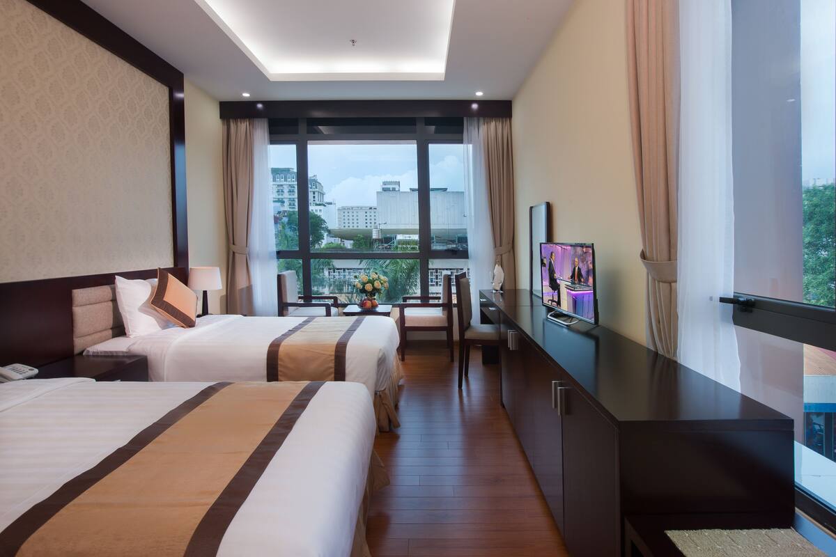 A hotel room with two beds, a desk, a TV, and a large window with a city view.