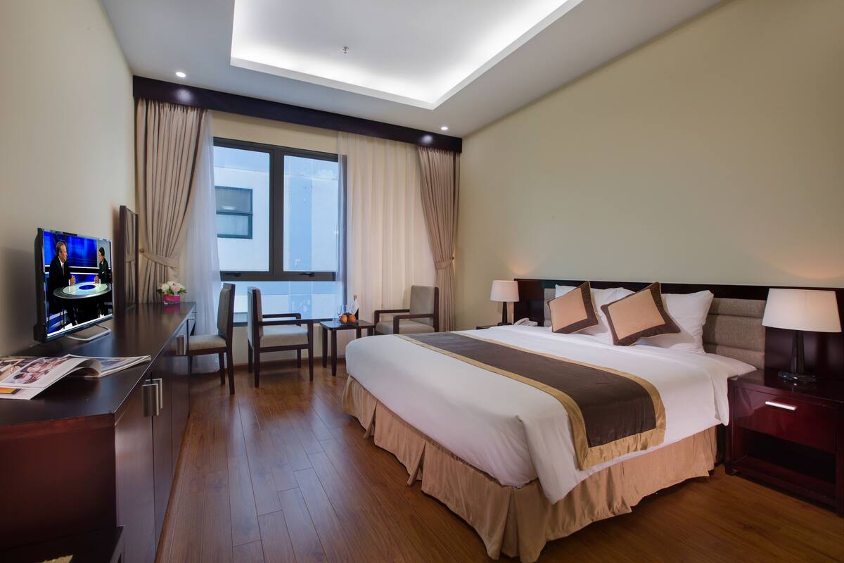 A hotel room with a large bed, a desk with a computer, a chair, a small table with a vase, and a window with curtains.
