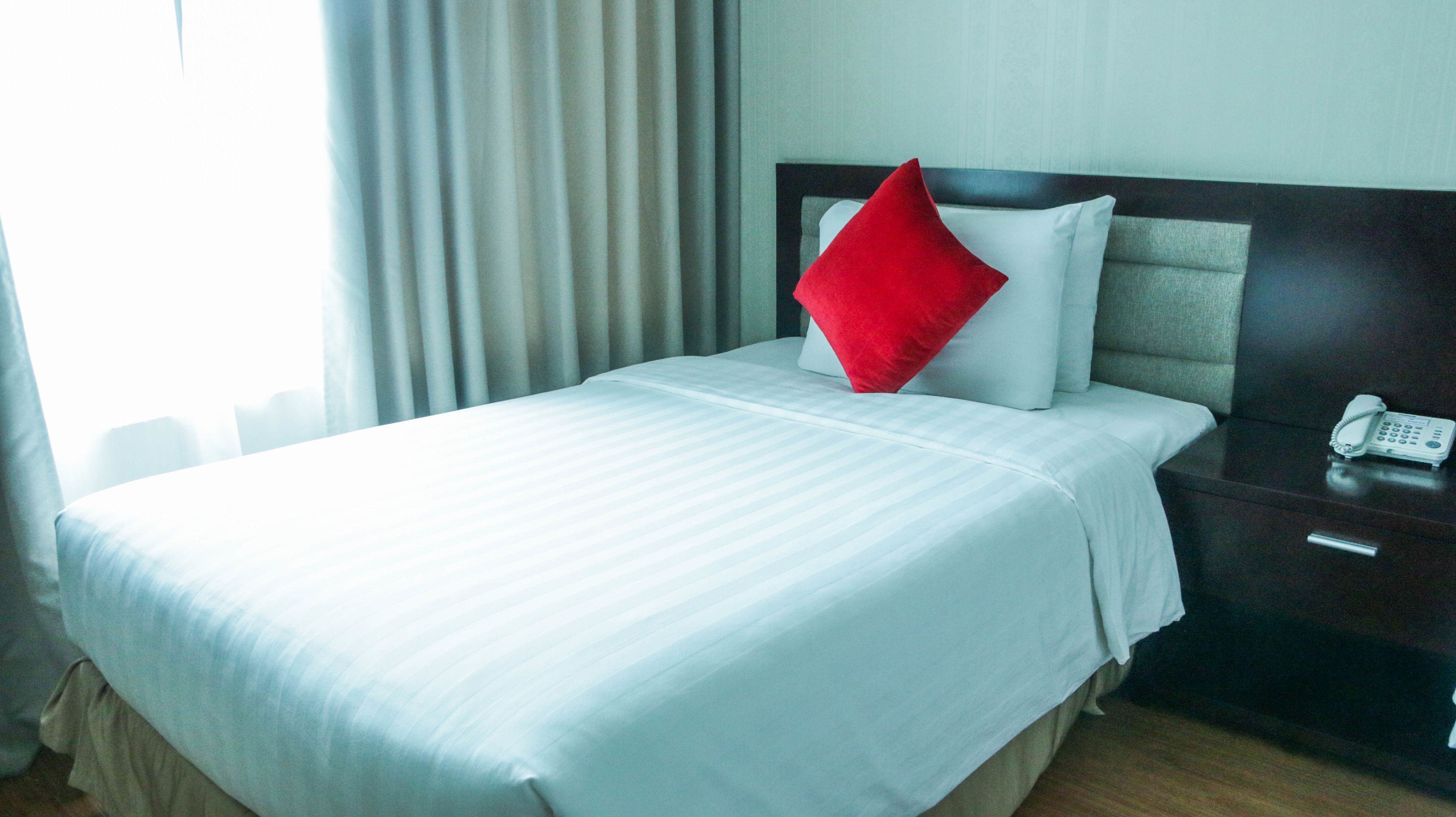 Ein Hotelzimmer mit einem ordentlich bezogenen Bett, einem Nachttischtelefon und einem Fenster mit Vorhängen.