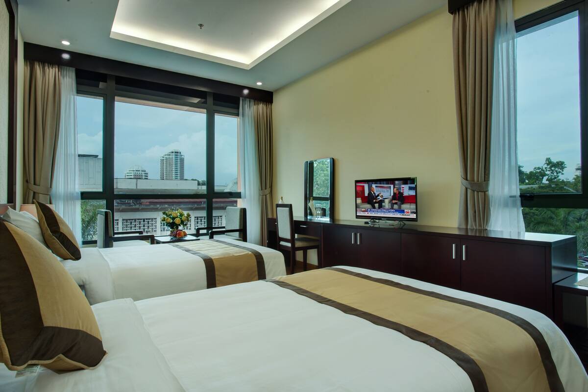 A hotel room with two beds, a desk, a TV, and a large window with a city view.