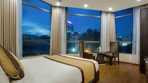 Une chambre d’hôtel avec un grand lit, un bureau et une vue sur le paysage urbain illuminé la nuit.