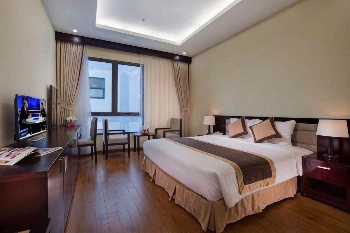 A hotel room with a large bed, a desk with a TV, a chair, a window with curtains, and a nightstand with a lamp.