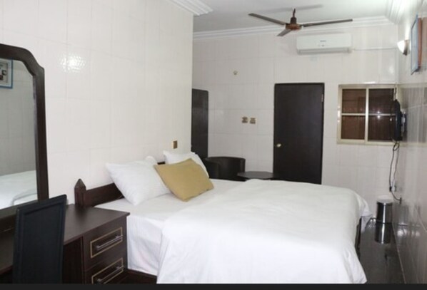 A hotel room with a bed, a desk, a chair, a wardrobe, a mirror, a ceiling fan, an air conditioning unit, a door, and a window.