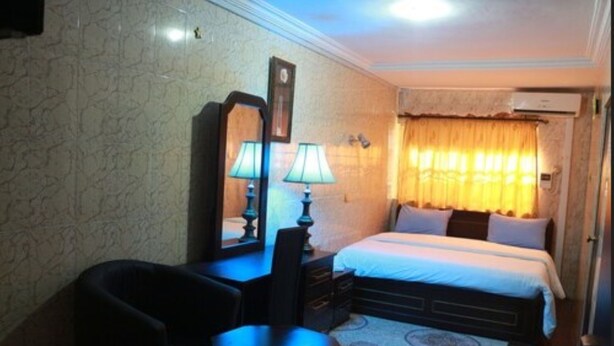 A hotel room with a bed, a desk with a chair, a mirror, and a lamp. - Cute Villa Hotel and Suites (Uyo)