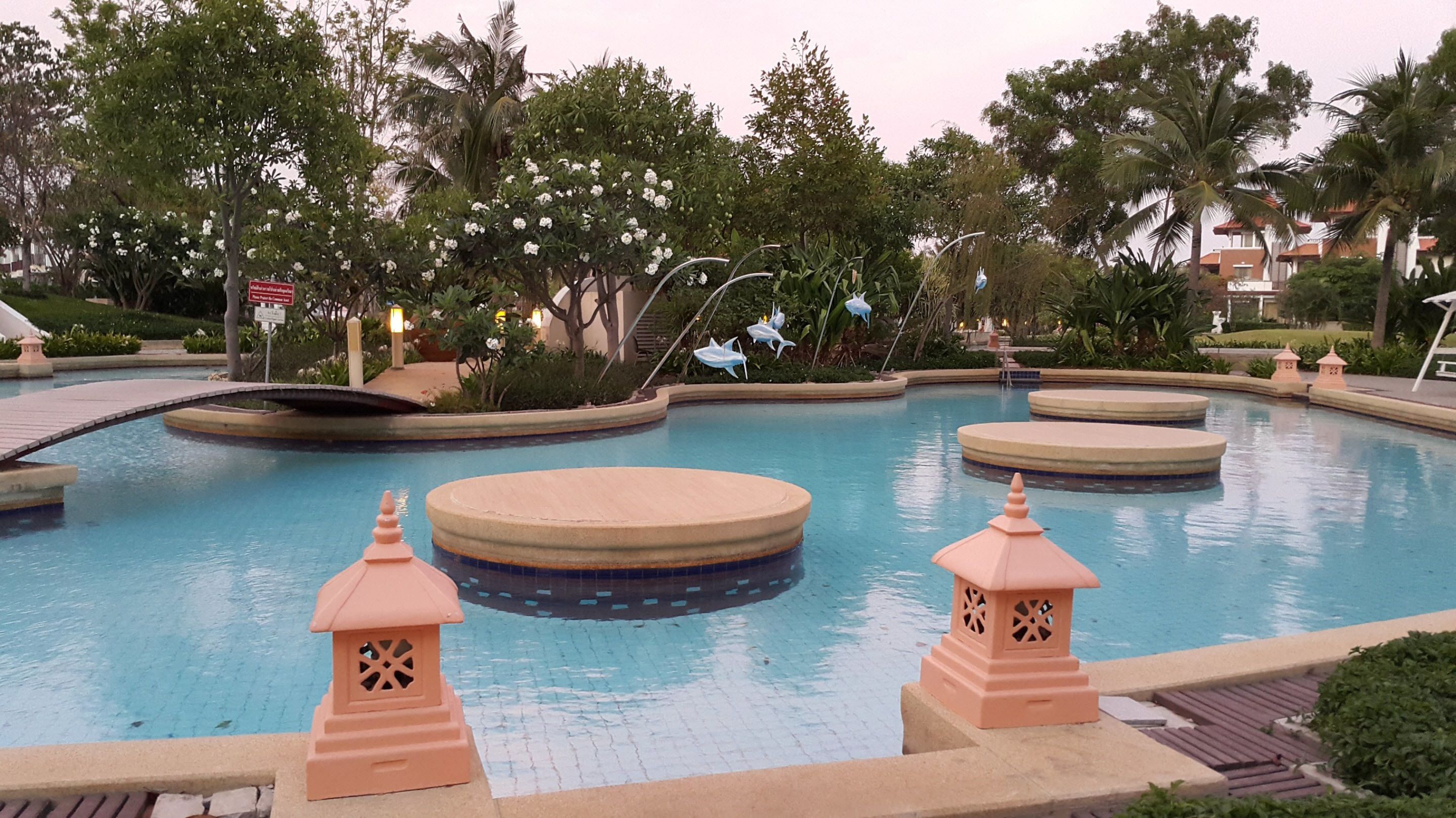 A swimming pool with a bridge, surrounded by lush greenery and palm trees.