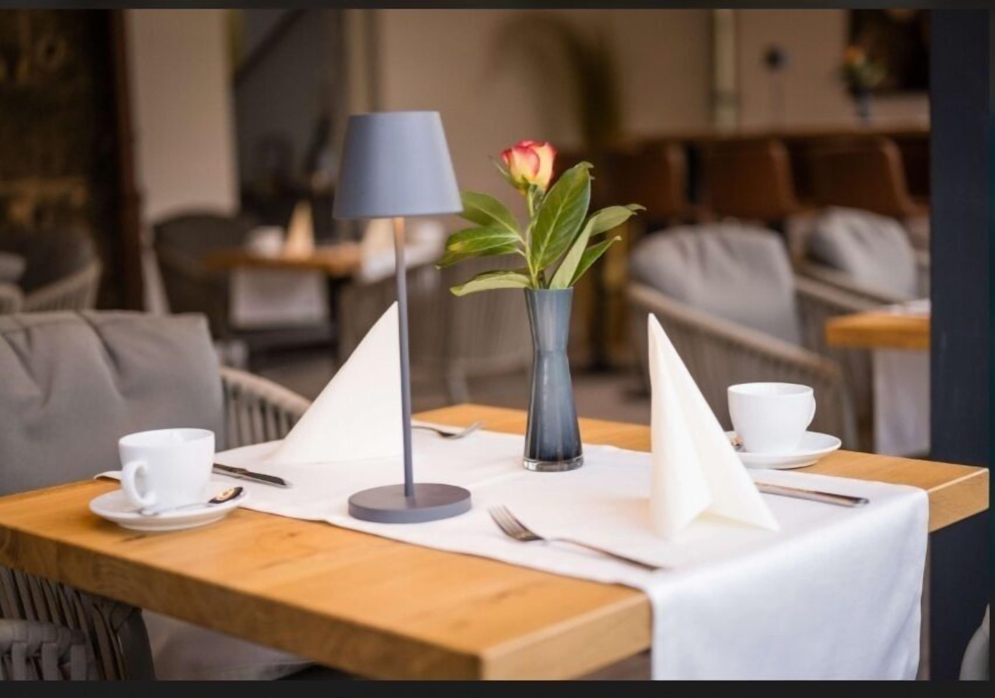 Una mesa vestida con mantel blanco, un florero con una sola flor, una lámpara, y una taza y platillo.