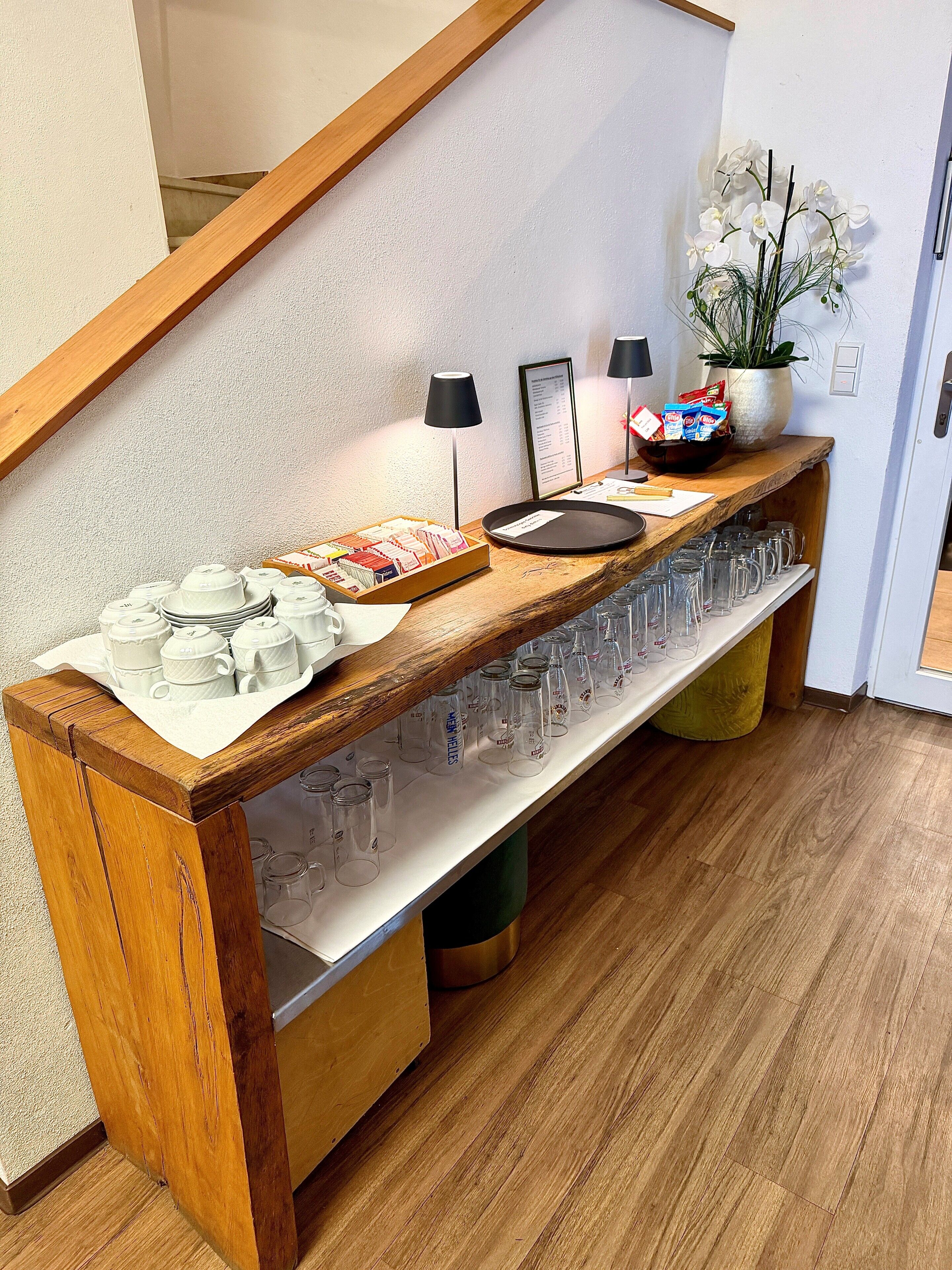 A wooden buffet table with a variety of food items, a potted plant, and a selection of glasses.