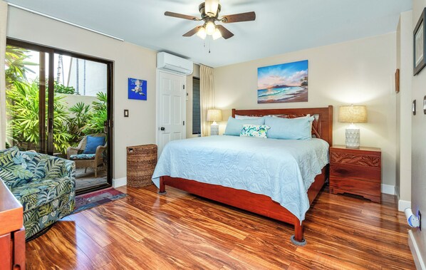 Un dormitorio con piso de madera, una cama con cubrecama azul, un ventilador de techo, una mesita de noche con lámpara y vistas al exterior a través de una puerta corrediza.
