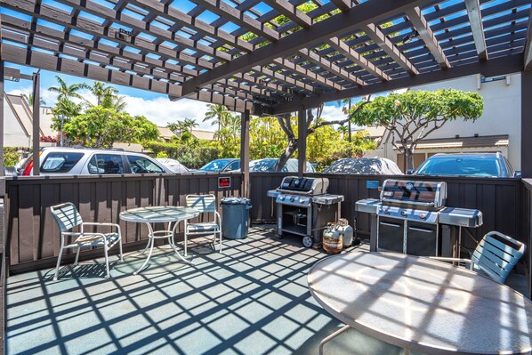 An outdoor patio area with a pergola, seating, a grill, and a view of parked cars and palm trees.