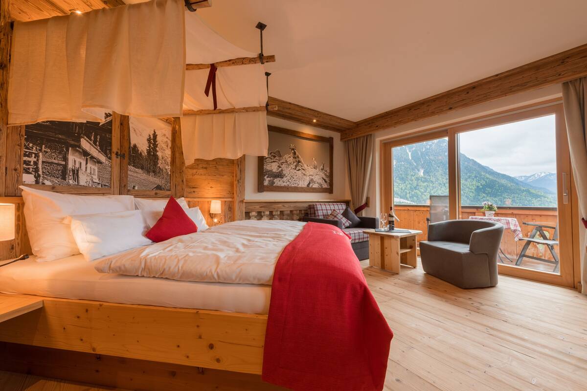 A wooden bed with a red blanket, a small table, a chair, and a large window with a mountain view.
