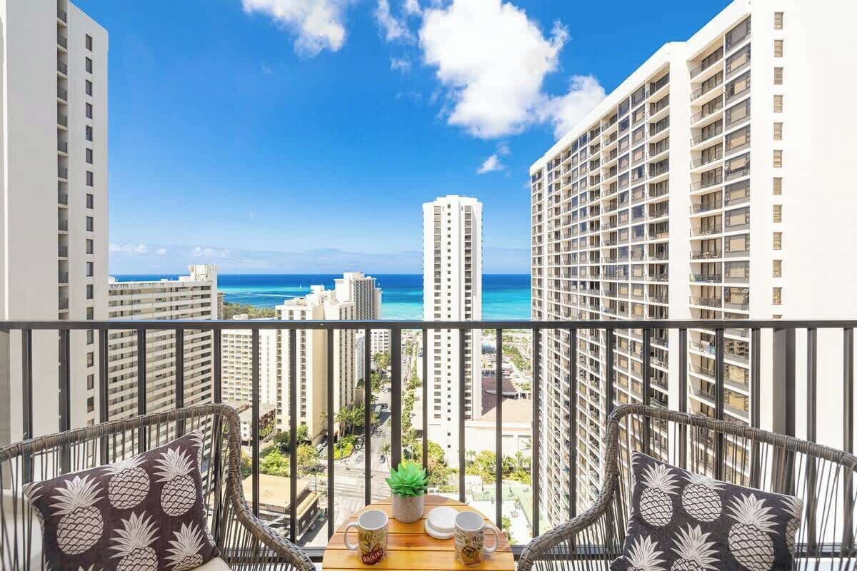 A balcony with wicker chairs, a small table with a plant, and a view of the ocean and high-rise buildings.