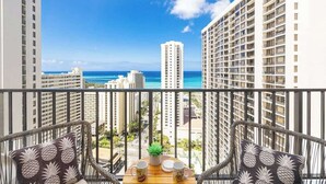 Ein Balkon mit Korbstühlen, einem kleinen Tisch mit Pflanze und Blick auf das Meer und hohe Gebäude.
