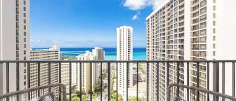 A balcony with wicker chairs, a small table with a plant, and a view of the ocean and high-rise buildings.