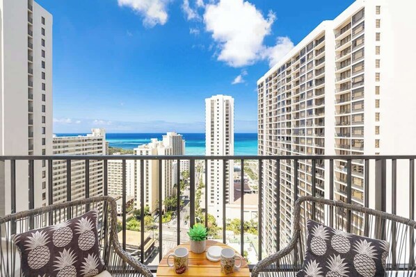 A balcony with wicker chairs, a small table with a plant, and a view of the ocean and high-rise buildings.