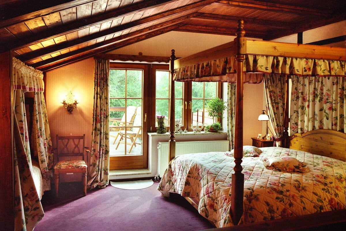 Ein Schlafzimmer mit einem Himmelbett, Holzboden, einem Fenster mit Blick auf Grünflächen, einem Sessel und einem Nachttisch mit Lampe.