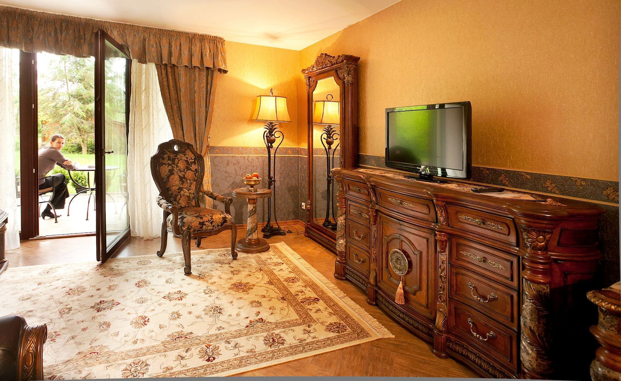 Ein Zimmer mit einem großen Spiegel, einem Fernseher, einem blumigen Ohrensessel und einem Tisch mit einer Lampe.
