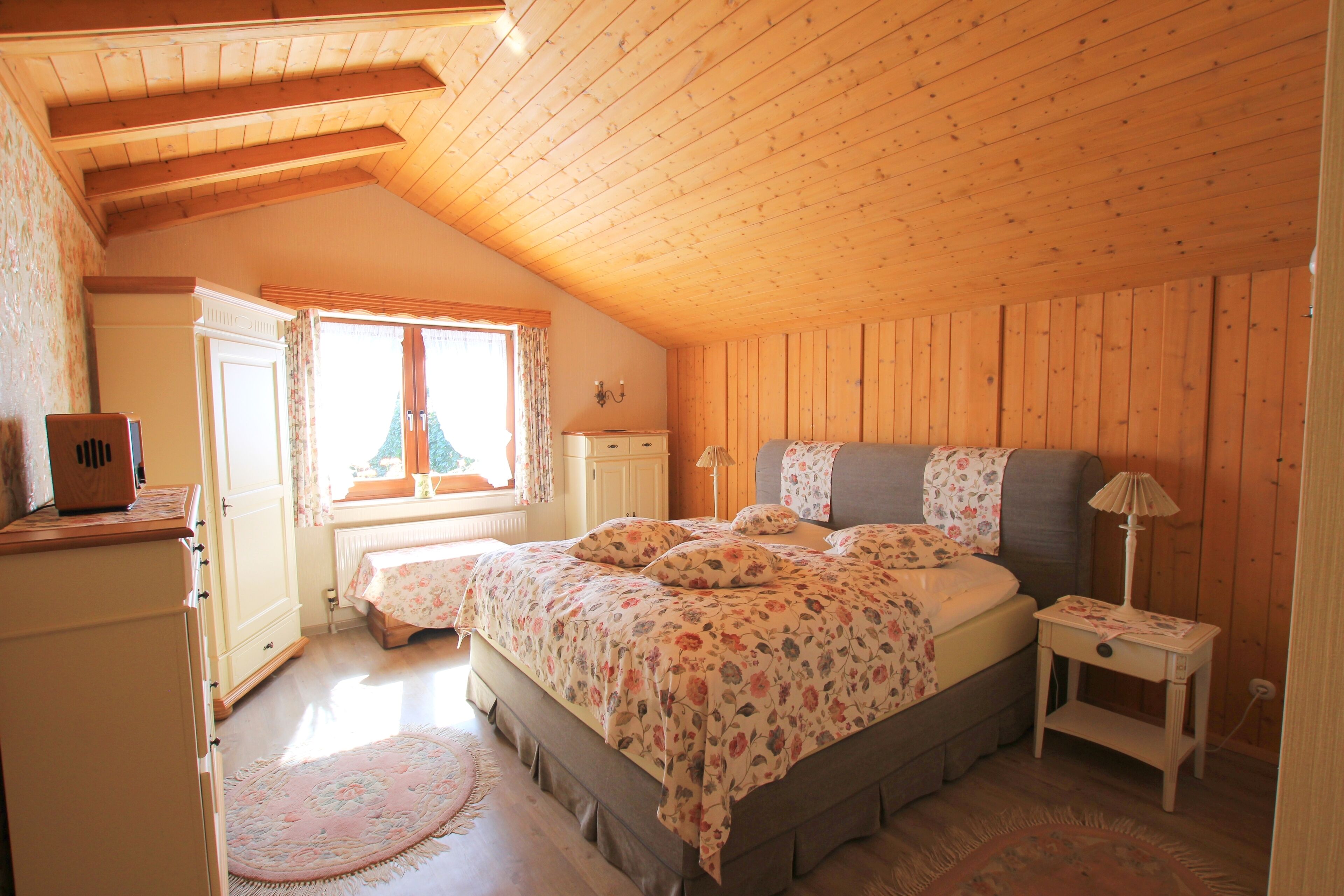 Un dormitorio con techo de madera, una cama con ropa de cama floral, una mesita de noche y un aparador.