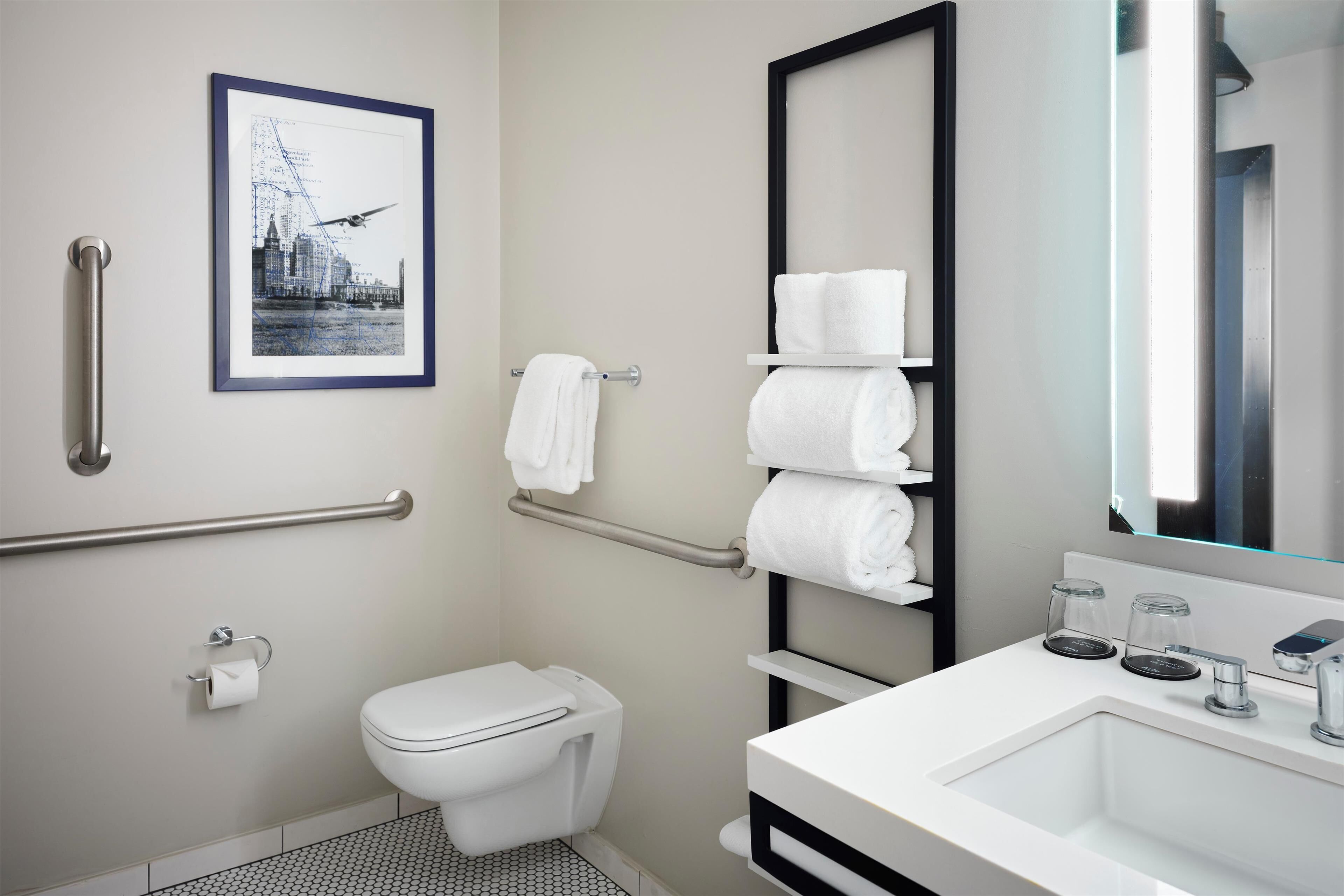 Un baño moderno con inodoro, barras de apoyo, lavabo con espejo y un cuadro en la pared.