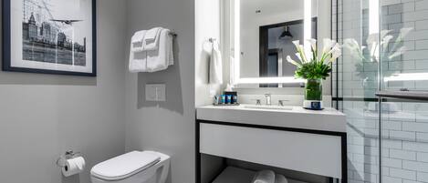 A modern bathroom with a toilet, sink, and mirror. There is a framed picture on the wall, a vase with flowers, and a towel rack.