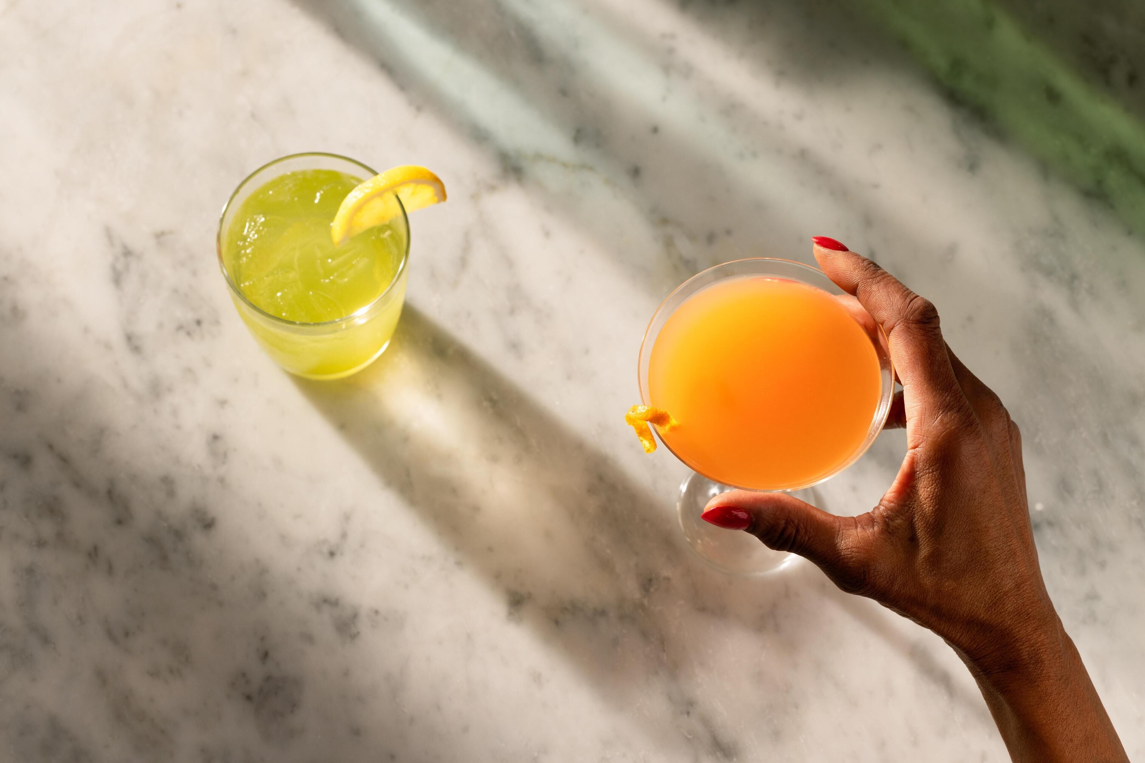Una mano sostiene dos vasos con bebidas, uno con un líquido amarillo y otro con un líquido anaranjado, sobre una superficie de mármol.