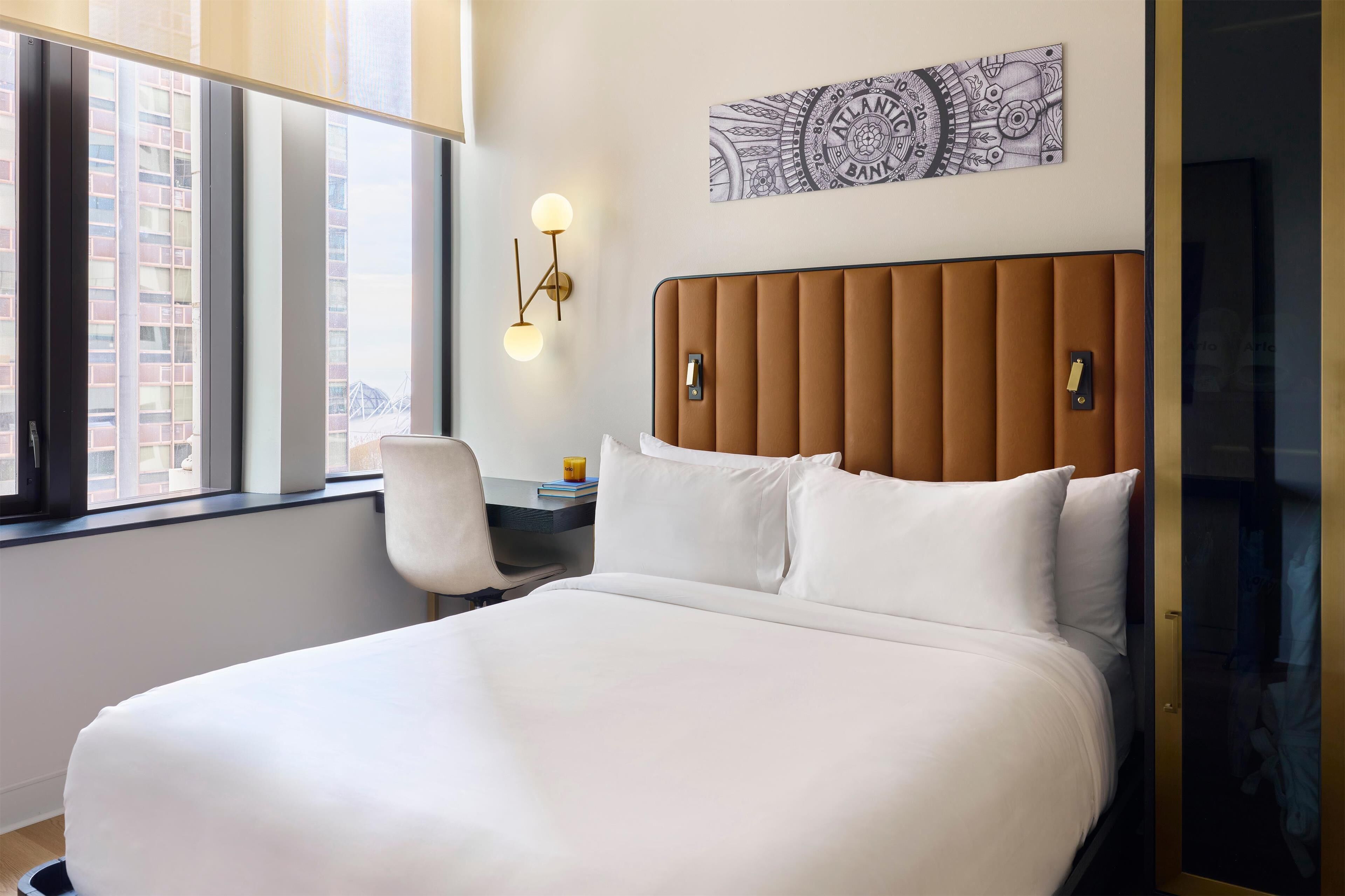 Habitación de hotel con una cama grande, un escritorio con silla, una ventana con vista a edificios y una obra de arte colgada en la pared.
