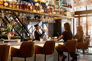 A bar area with a bartender, two seated customers, and a variety of liquor bottles on shelves. - Arlo Chicago (Chicago)