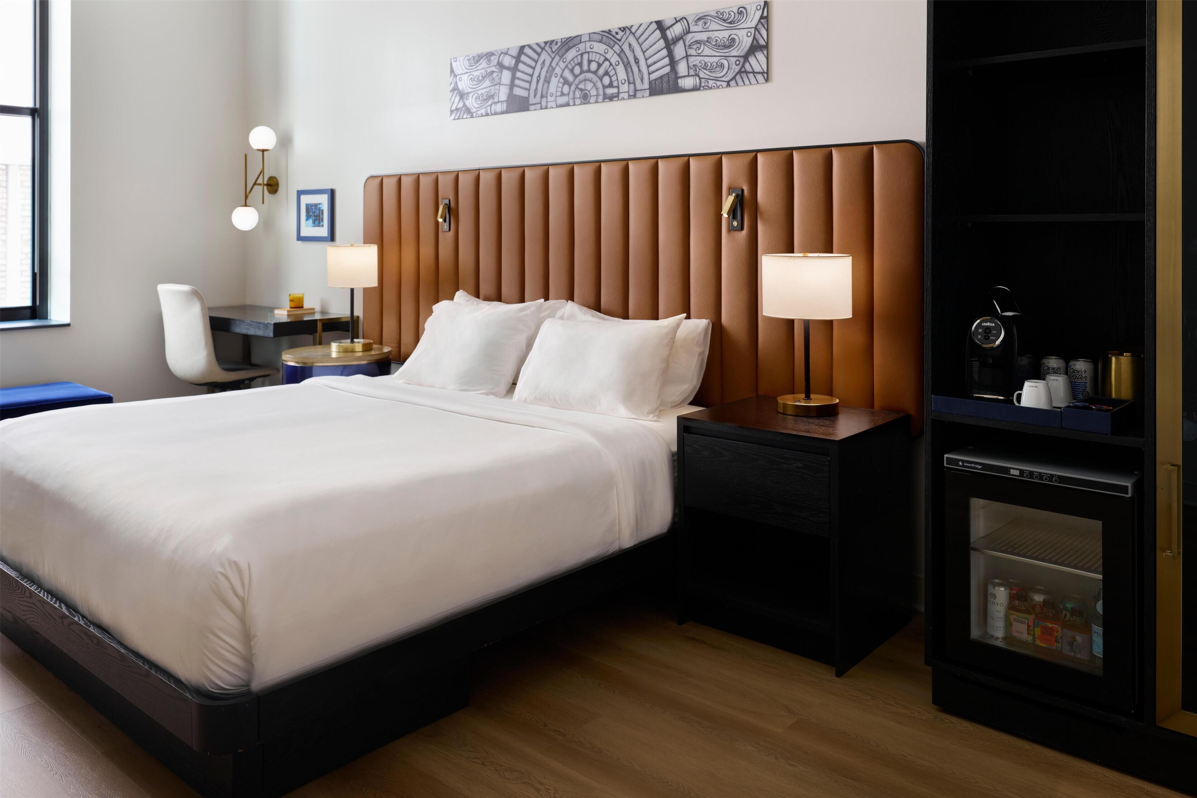 Una habitación de hotel con una cama grande, dos mesitas de noche, un escritorio, una silla, un armario y una obra de arte colgada en la pared.